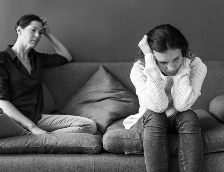 Psycholog online. Czarno-białe zdjęcie przedstawiające dwie smutne, strapione kobiety (matkę i córkę) siedzące na kanapie, odwrócone od siebie. Córka (z przodu, w jasnym swetrze i dżinsach) obejmuje rękami głowę, patrząc w dół. Matka (z tyłu, w ciemnej koszuli) podpiera głowę ręką, wpatrzona w dal. Kompozycja boczna, oświetlenie studyjne.