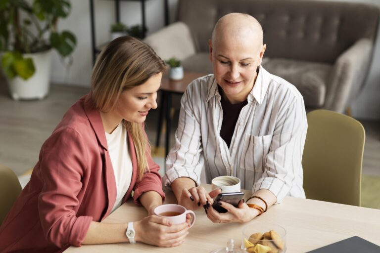Kobieta w trakcie leczenia onkologicznego i jej bliska osoba siedzą przy stole, wspólnie korzystając z telefonu. Zdjęcie ilustruje wsparcie bliskich i konsultacje psychologiczne online w chorobie nowotworowej.