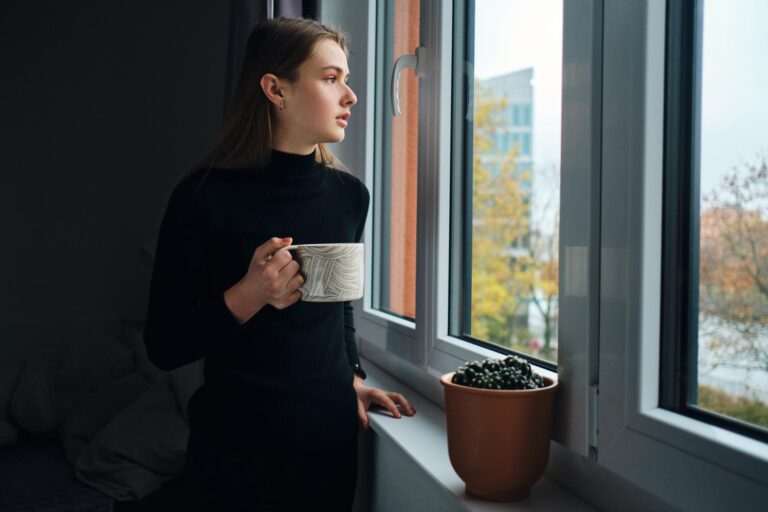 Kobieta wpatrzona w okno w pochmurny jesienny dzień, czując się przygnębiona.